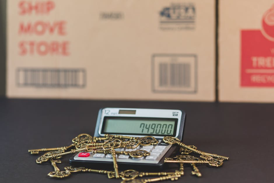 A close-up view of a small calculator displaying the number 749,800, surrounded by a scattered collection of gold-colored keys of various sizes and styles. The calculator is situated on a dark surface inside a property, with several cardboard boxes in the background, some labeled with red and black text related to moving or shipping. The scene captures a moment during packing or preparations for a home relocation, with the keys possibly representing furniture or apartment keys being organized before a move. The background boxes are partially out of focus, emphasizing the keys and calculator in the foreground, indicating an organized approach to moving logistics, as managed by Man With a Van Hampstead, a service specializing in removals.