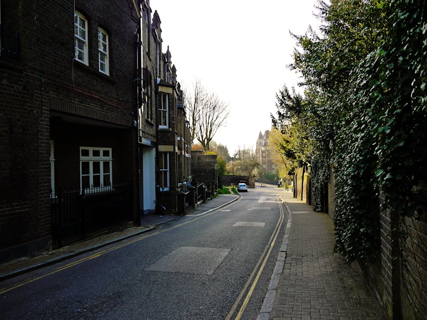 A quiet residential street in Hampstead with a narrow tarmac road featuring double yellow lines along the curb. On the left side, there are brick terraced houses with white-framed windows, some with small front gardens enclosed by black metal fences. On the right, a stone-paved sidewalk runs parallel to a tall, dense hedge that overhangs onto the pavement. The scene is illuminated by soft natural light during daylight hours, with leafless trees casting shadows across the street. In the distance, a white van is parked on the street, and a building with a tower and spire can be seen further along the road. The image captures the typical environment where home relocation and furniture transport operations, such as loading and unloading belongings, are conducted, often involving the use of vans and packing materials. The presence of a white vehicle and the edge of a moving van suggest ongoing or upcoming loading activities, with interior items like cardboard boxes or furniture possibly being prepared for transport later in the process. Man With a Van Hampstead’s services are sometimes involved in such residential moving scenarios.