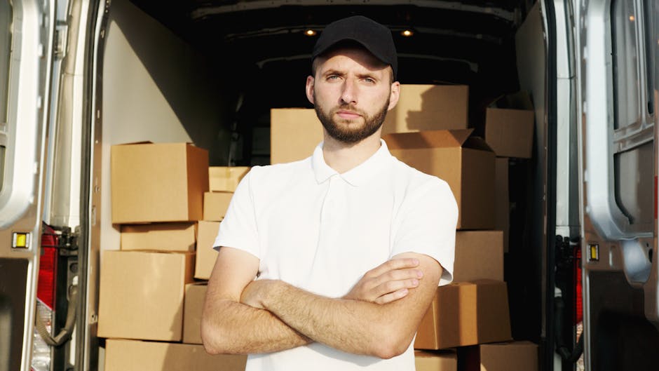 A young man with a beard and serious expression, wearing a white polo shirt and black cap, stands inside the open rear of a moving van. Behind him, several large cardboard boxes are stacked on the van