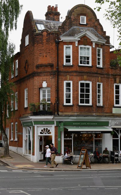 A three-story red brick building with a curved parapet roof and multiple white-framed sash windows is situated on a street. The ground floor houses a bakery, café, and patisserie with a green and white facade, including an entrance with a decorative arched door and large display windows. In front of the shop, several people are seated at outdoor tables on the pavement, and a few pedestrians are walking past. The building is adjacent to a sidewalk lined with trees, and a portion of the street with tram or bus tracks is visible in the foreground. This scene depicts a typical urban setting that could be part of a house removals or relocation process, with the building's ground floor and surrounding environment clearly visible for context, supported by Man With a Van Hampstead's professional services.