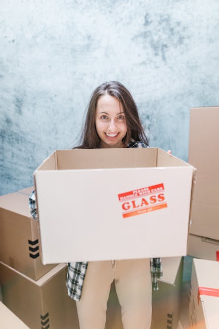 A young woman with long brown hair smiling while holding a large empty cardboard box labeled 'Fragile - Handle with Care - Glass' in an indoor setting. She is surrounded by several other packed cardboard boxes of various sizes, some sealed with packing tape, and standing in front of a textured light blue wall. The scene depicts the packing stage of a home relocation, with the woman preparing for a move managed by a professional removals service. The boxes are arranged on the floor and nearby surfaces, indicating an organized packing process. This image is relevant to house removals and moving logistics, illustrating the packing aspect of furniture transport and home relocation.