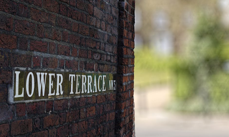 Close-up view of a brick wall with a metal street name sign reading 'Lower Terrace NW3' affixed to the left side of the image. The sign features white and green lettering on a silver background. To the right, the image shows a blurred outdoor scene with greenery and a pathway, suggesting an exterior residential area in Hampstead. Inside the property, partially visible are packed cardboard boxes, some wrapped in protective plastic film or blankets, placed on the floor near a doorway. The scene hints at the process of home relocation, with furniture and boxes prepared for loading into a van. Visible are a green moving strap and a trolley or dolly used for transporting household items, emphasizing the logistics involved in furniture transport and packing during a house move. The lighting is natural, indicating daytime, and the setting is calm and organized, consistent with professional removal services like those offered by Man With a Van Hampstead.