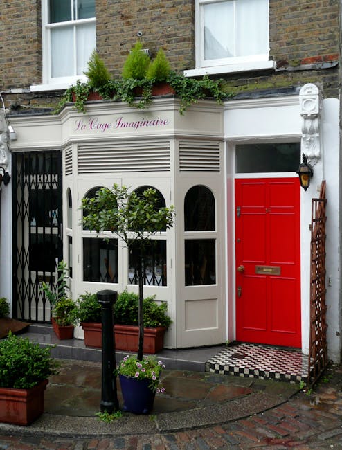 The image shows the exterior of a shop located on Hampstead High Street, featuring a traditional brick building with two white-framed windows on the second floor. Below the windows, there is a charming white shopfront with a curved, wooden facade and a sign reading 'La Cage Imaginare' above it. In front of the shop, on the pavement, there are several potted plants including small trees and flowering plants in terracotta and plastic containers, enhancing the entrance's visual appeal. To the right of the shopfront, there is a bold red-painted door with a mail slot and a black lantern-style light fixture mounted beside it. Adjacent to the door is a wooden lattice panel leaning against the wall. The scene appears set during daylight with overcast sky, suitable for house removals or furniture transport prep, and the location is associated with Man With a Van Hampstead’s local removals and packing services, highlighted by the clean, organized setting, which could relate to a home relocation or moving process.