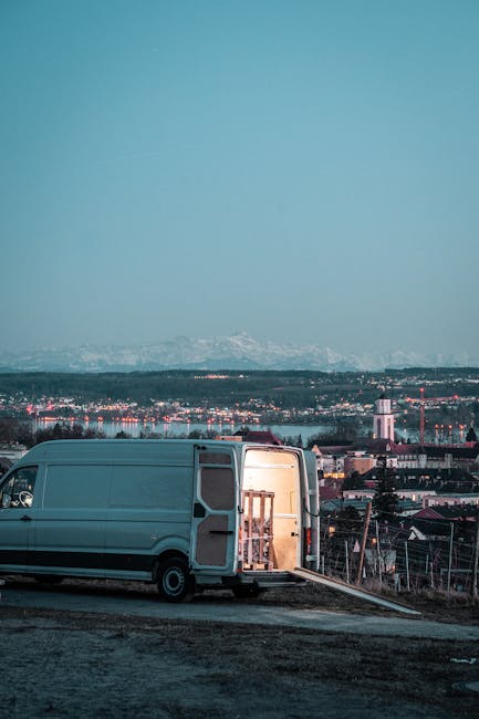 A large white moving van with its side door open, revealing a wooden furniture piece inside, parked on a paved roadway in an elevated area overlooking the cityscape of Hampstead, with residential buildings, rooftops, and a church steeple visible in the background, under a clear blue sky. The van is positioned near the edge of the pavement, suggesting the initial stages of a home relocation or furniture transport process, with the open door facilitating the loading or unloading of household items. The surrounding environment is a mix of natural and urban elements, and the scene is lit by natural daylight, emphasizing the practical aspect of house removals conducted by Man With a Van Hampstead, specializing in professional moving services including packing, loading, and transporting household furniture and belongings in the Hampstead and Belsize Park areas.