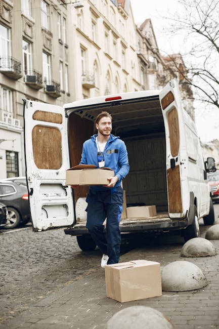 A large white moving van with its side door open, revealing a wooden furniture piece inside, parked on a paved roadway in an elevated area overlooking the cityscape of Hampstead, with residential buildings, rooftops, and a church steeple visible in the background, under a clear blue sky. The van is positioned near the edge of the pavement, suggesting the initial stages of a home relocation or furniture transport process, with the open door facilitating the loading or unloading of household items. The surrounding environment is a mix of natural and urban elements, and the scene is lit by natural daylight, emphasizing the practical aspect of house removals conducted by Man With a Van Hampstead, specializing in professional moving services including packing, loading, and transporting household furniture and belongings in the Hampstead and Belsize Park areas.