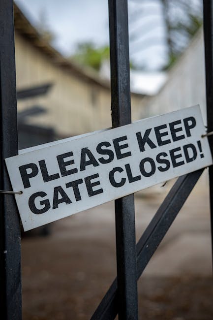 Close-up of a metal gate with a white sign attached to it, displaying the message 'Please Keep Gate Closed!' in bold black letters. The sign is fastened with metal hooks on a black-painted gate, which